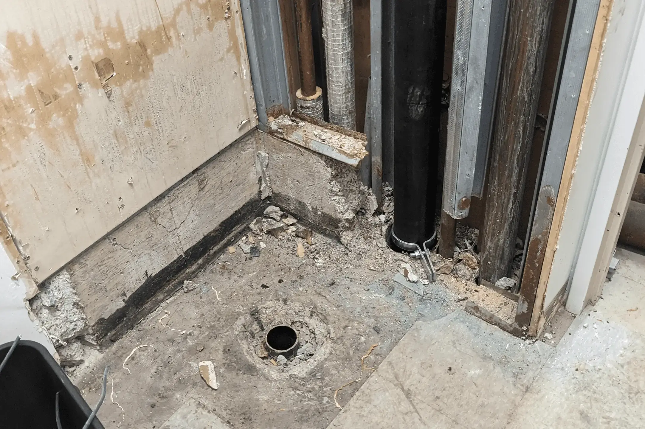 Exposed pipes and a floor drain in a corner of a room under renovation.